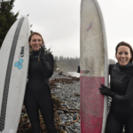 Two women stand with surf boards