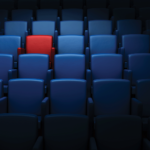 A movie theatre full of blue seat with one red seat standing out on the top left corner