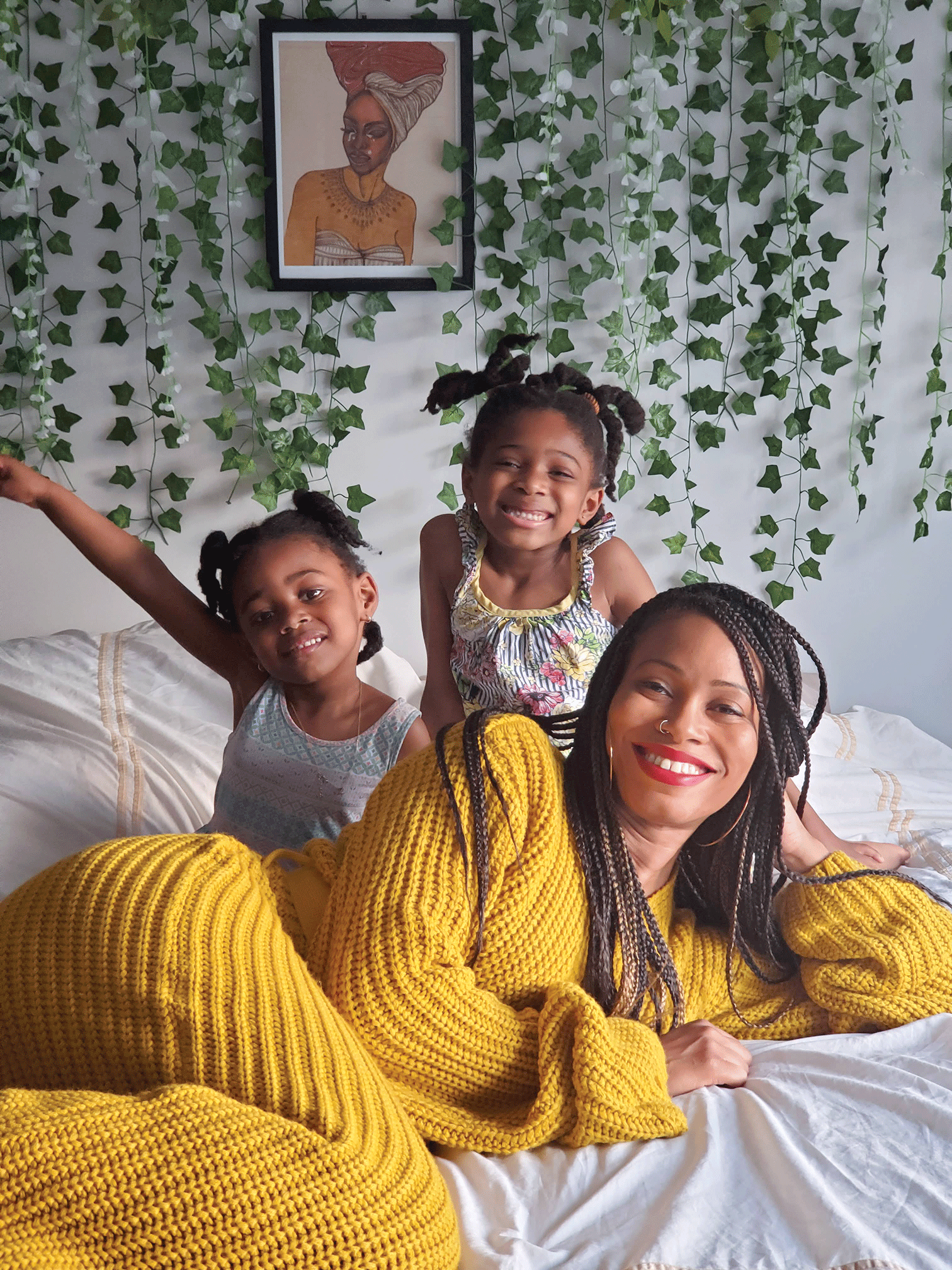 Mother and two young daughters smiling while laying on a bed with greenery in the background.