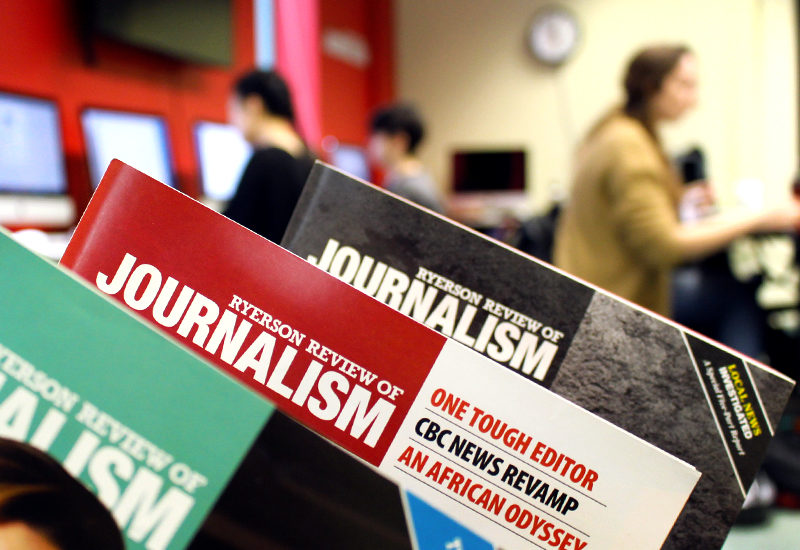 Closeup of three Review of Journalism magazines with students working on computers in the background