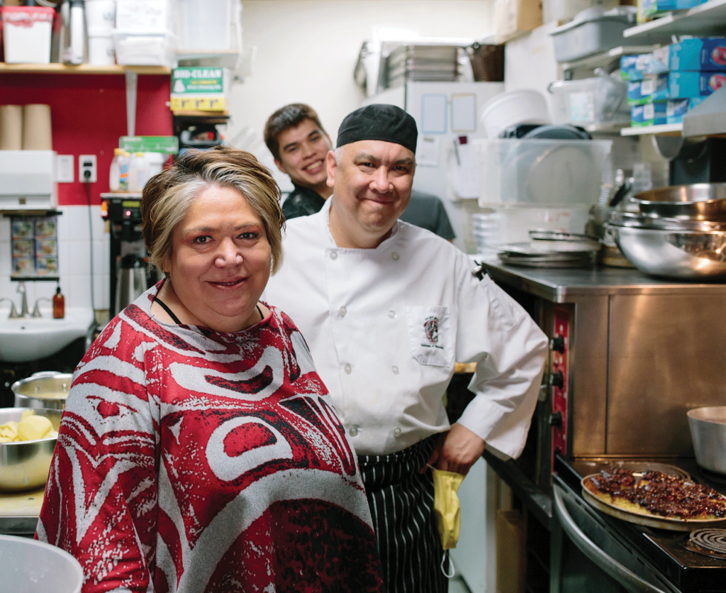 Three people including a chef standing in a cramped kitchen next to a stove