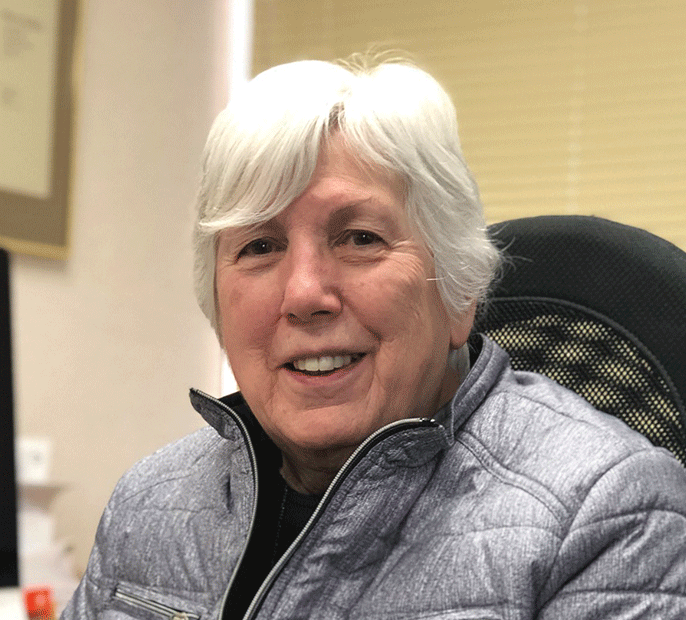 A woman with white hair in a jacket sitting in an office.