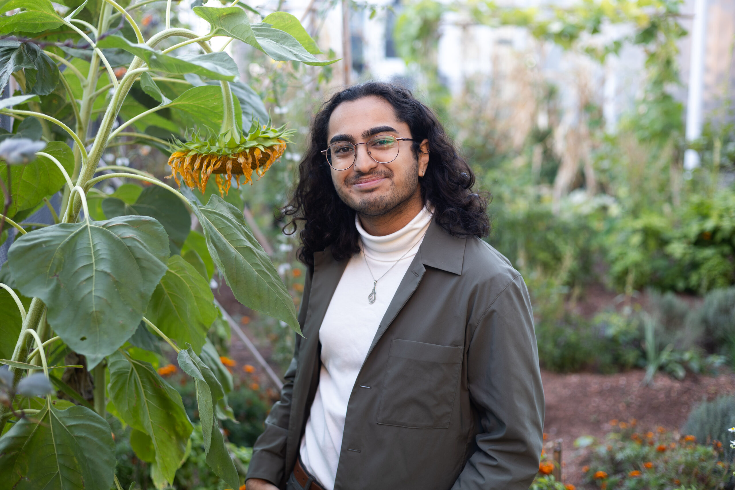 Aliyaan Amlani-Kurji is pictured smiling wearing a white turtleneck with a gray blazer, and glasses. To his left is a sunflower drooping down with large dark green leaves.