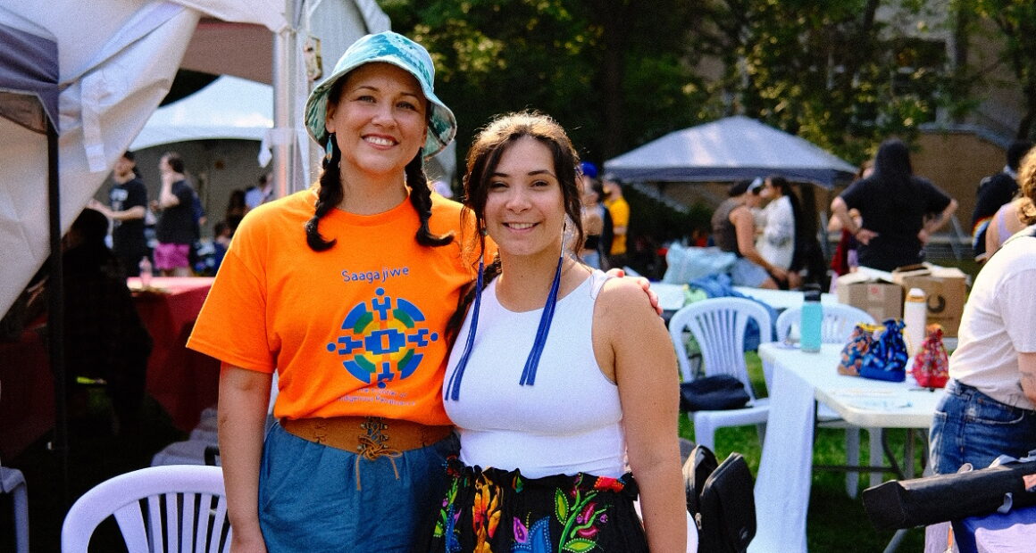 Gabby McMann (right) and Carrie Davis (left) are hip-hip. McMann wears a white tank top and a floral satin skirt. The colors are pink, magenta, black, blue, orange and green. The skirt has embroidered flowers and leaves with green and pink, and larger blue tulip petals with a pink flower bud. To the left, Davis wears an orange Saagajiwe Indigenous Studio orange t-shirt that has a blue, green, orange, logo shaped symmetrically in an embroidered stitched pattern in the centre.