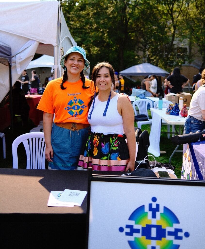 Gabby McMann (right) and Carrie Davis (left) are hip-hip. McMann wears a white tank top and a floral satin skirt. The colors are pink, magenta, black, blue, orange and green. The skirt has embroidered flowers and leaves with green and pink, and larger blue tulip petals with a pink flower bud.  To the left, Davis wears an orange Saagajiwe Indigenous Studio orange t-shirt that has a blue, green, orange, logo shaped symmetrically in an embroidered stitched pattern in the centre. 
