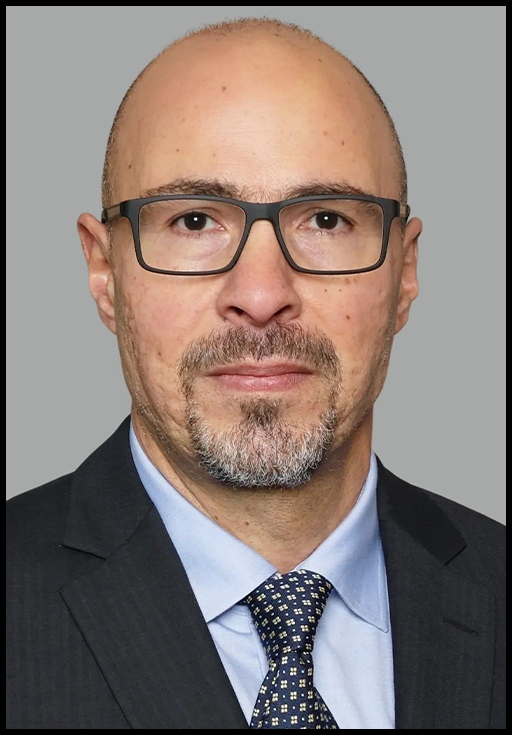 A bald man with a beard wearing glasses and a suit and tie posed in front of a grey background.