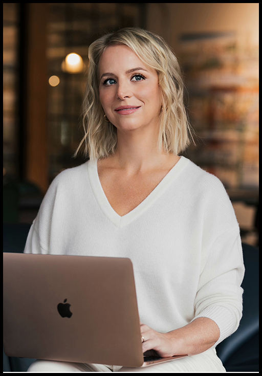 A woman with short blonde hair wearing a white sweater sitting and holding a computer in front of a blurred, dim lit background.