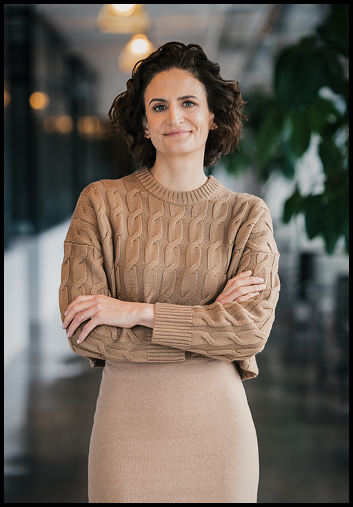 A woman with short brown hair wearing a beige sweater and skirt standing and crossing her arms in front of a blurred office background.