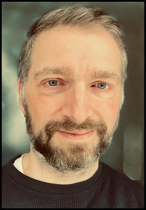 A man with a beard wearing a black shirt posed in front of a blurred background.