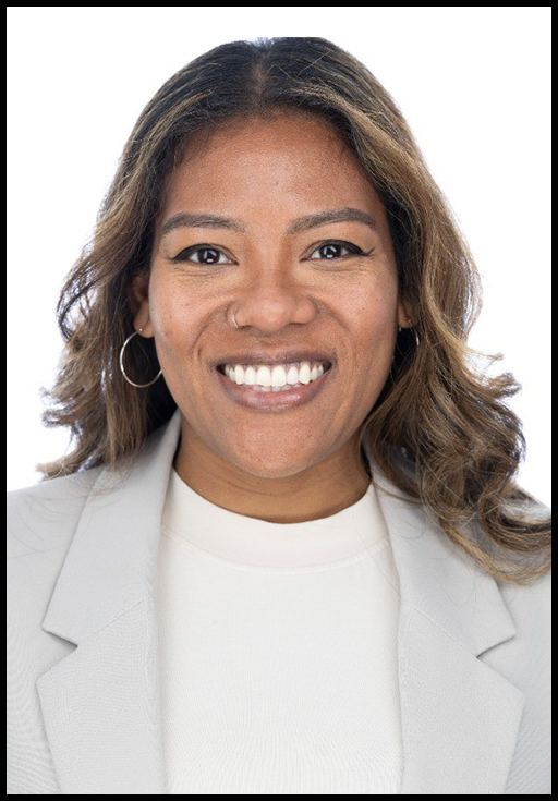 A Black woman with brown wavy hair and wearing a light grey blazer smiling in front of a white background.