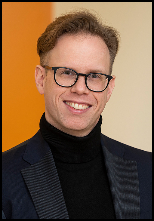 A man wearing glasses, a black blazer and a black turtleneck posed in front of an orange and beige background.