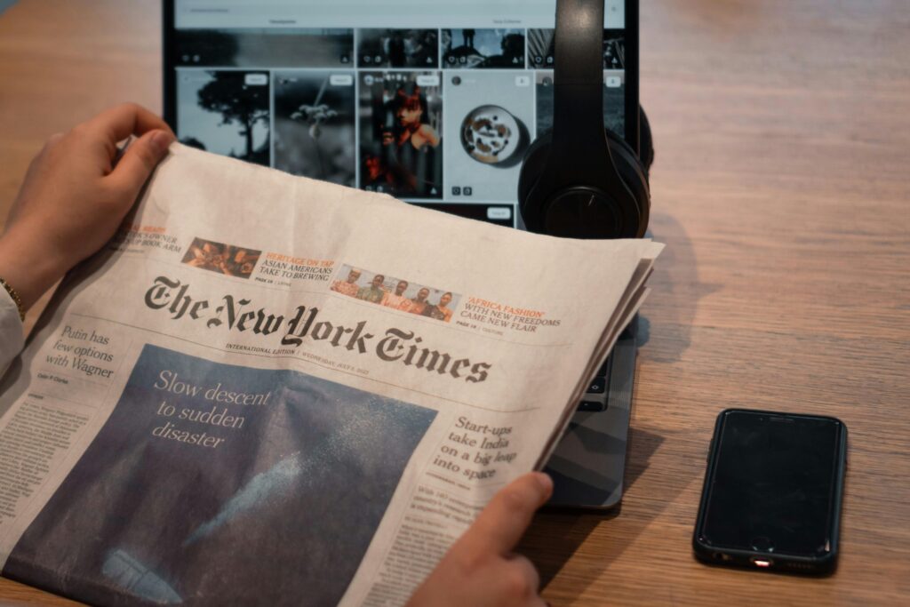 Two hands holding a newspaper in front of a computer with blurred out images and headphones hanging off the screen. A black smartphone is placed on a wooden table to the right of the computer.