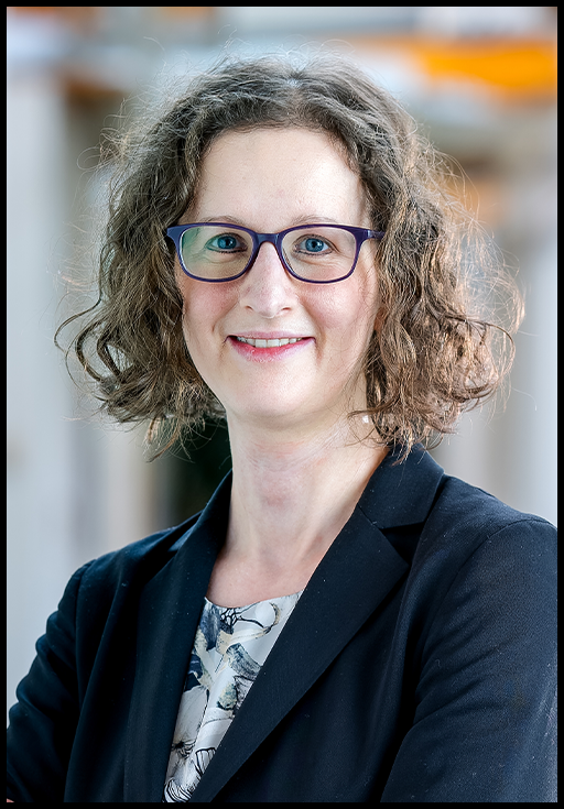 A woman with short, curly brown hair wearing glasses and a blazer posed in front of a blurred background.