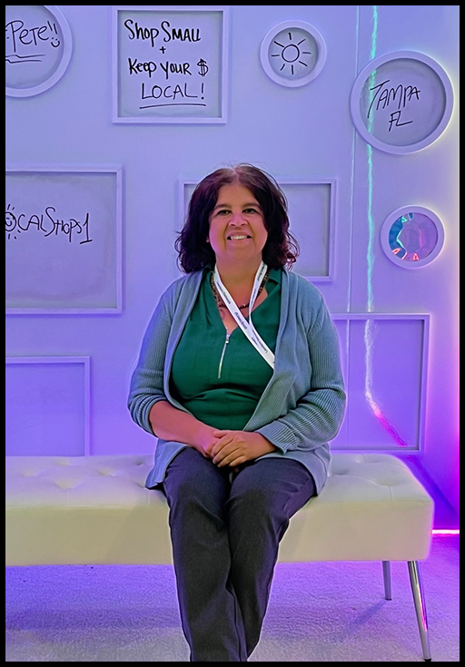 A woman with brown hair wearing a blue cardigan sitting on a white bench in front of a wall with white frames with words written inside