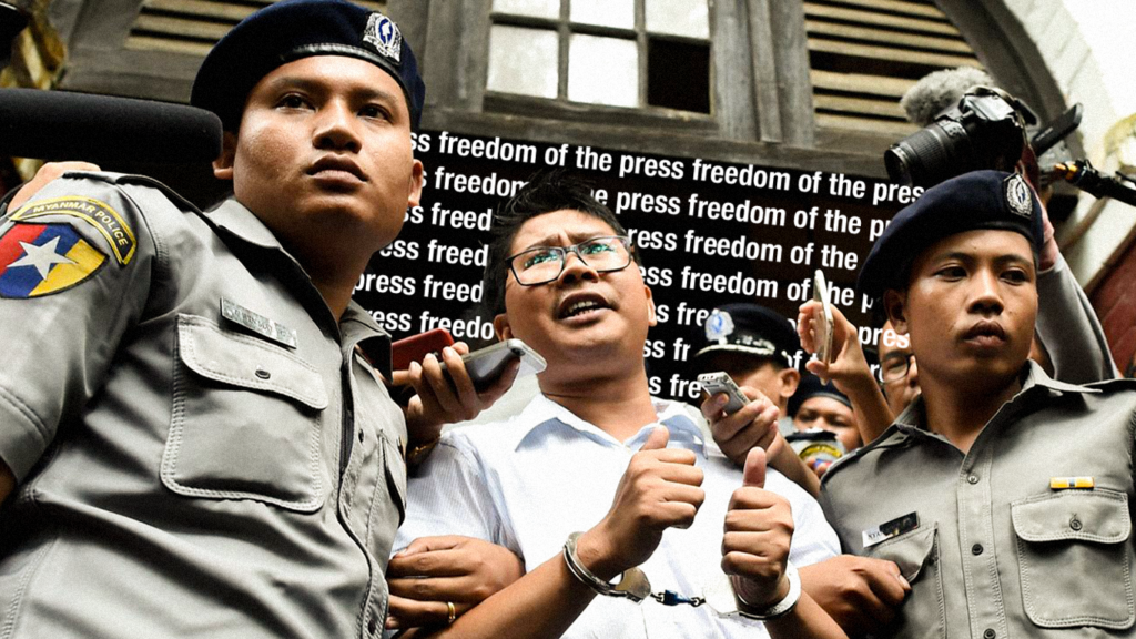 A person in a white shirt and glasses standing between two Myanmar police officers while wearing handcuffs. In the background reads, 