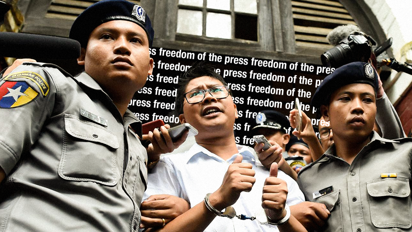 A person in a white shirt and glasses standing between two Myanmar police officers while wearing handcuffs. In the background reads, "Press Freedom" repeating across a screen in white letters.