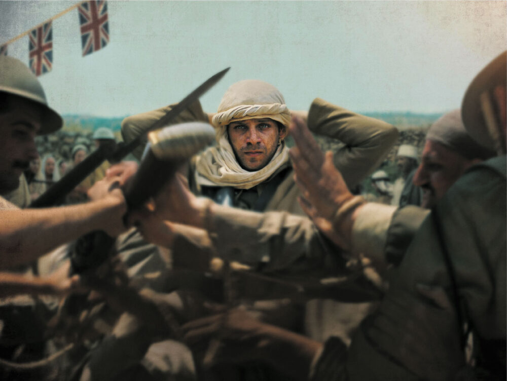 A colorized image depicts a man with striking blue eyes and a light-colored ghutra (head covering), appearing to be captured or struggling, as blurred figures surround him. His hands are near his head, and a blade is visible by his left shoulder. Partially visible British soldiers in helmets are to the left and right, with a blurred crowd behind. Two Union Jack flags hang against the light sky in the upper left, indicating a historical colonial setting.